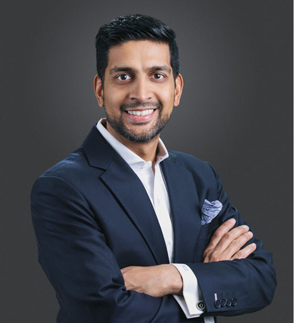 A smiling man in a business suit stands with his arms crossed.