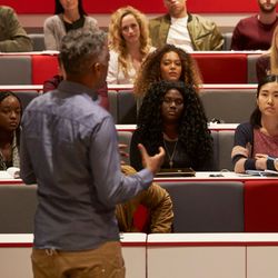 A professor, facing away from the camera, teaching a class of undergrads, facing the camera